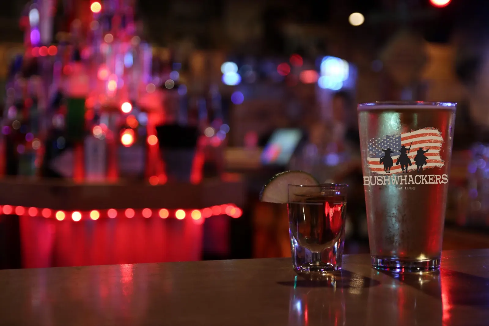 A Bushwhackers pint glass printed with the three-riders American flag logo beside a whiskey shot with a lime wedge, the bottle rail glowing red behind the bar